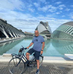 A man standing beside a bicycle in front of a modern architectural building with a pool, under a partly cloudy sky.