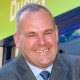 A man in a suit and tie, smiling, with a green and blue sign partially visible in the background.